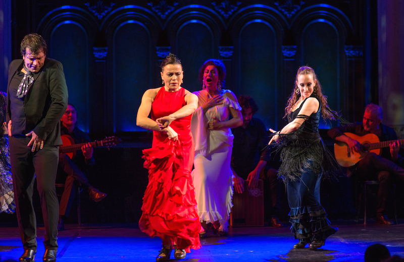 Soledad Barrio holds on to the ruffles of her bright red dress while a singer in white performs alongside a host of musicians. Two dancers in black flank Barrio.